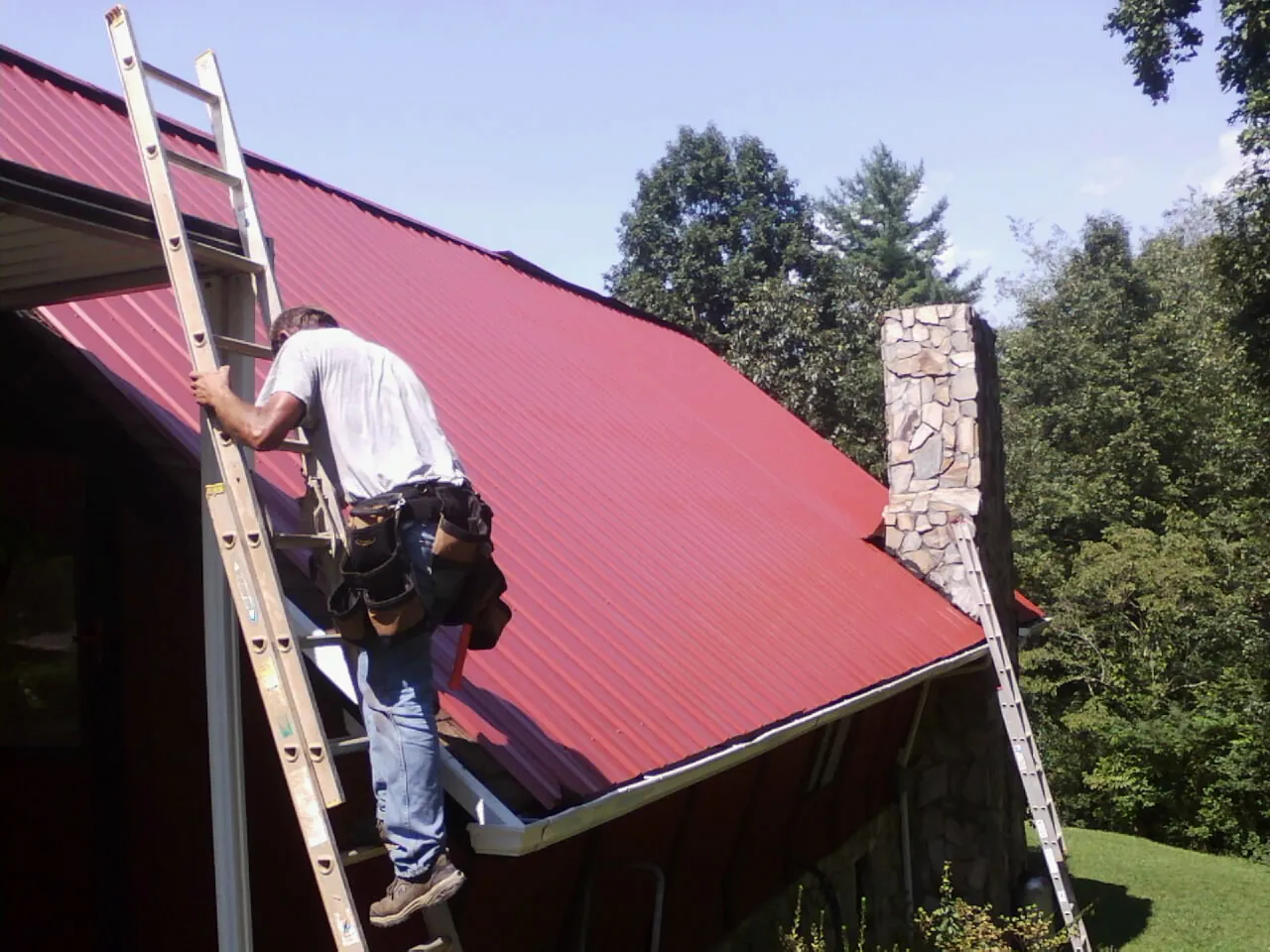 Roofer on a ladder beside a red metal roof and stone chimney — Soffit & Fascia Repair by Roofing Brothers in Wisconsin Rapids